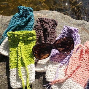 Może przedstawiać: Kolekcja ręcznie robionych szydełkowych etui na okulary w różnych kolorach, w tym turkusowym, brązowym, limonkowym, fioletowym, różowym i niebieskim. Każde etui ma białe dno i zamknięcie na sznurek. Okulary przeciwsłoneczne spoczywają na etui, wystawione na skale w pobliżu wody.