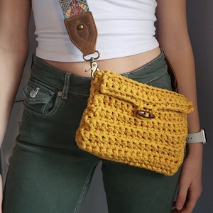May include: A yellow crocheted crossbody bag with a brown leather strap and a gold clasp. The bag is worn over a white shirt and green jeans.