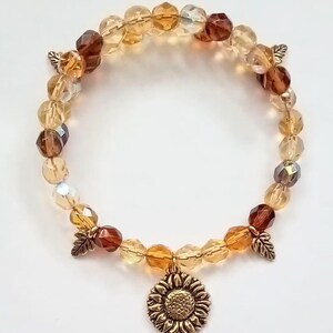 Könnte beinhalten: Ein braunes, goldenes und klares Glasperlenarmband mit einem Sonnenblumen-Anhänger. Das Armband hat kleine goldene Blattakzente.