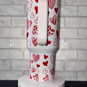 Puede incluir: Taza de viaje blanca con asa blanca y un patr&oacute;n de corazones rojos. La taza est&aacute; sobre una plataforma giratoria blanca con el texto "Magically Perfected Co."