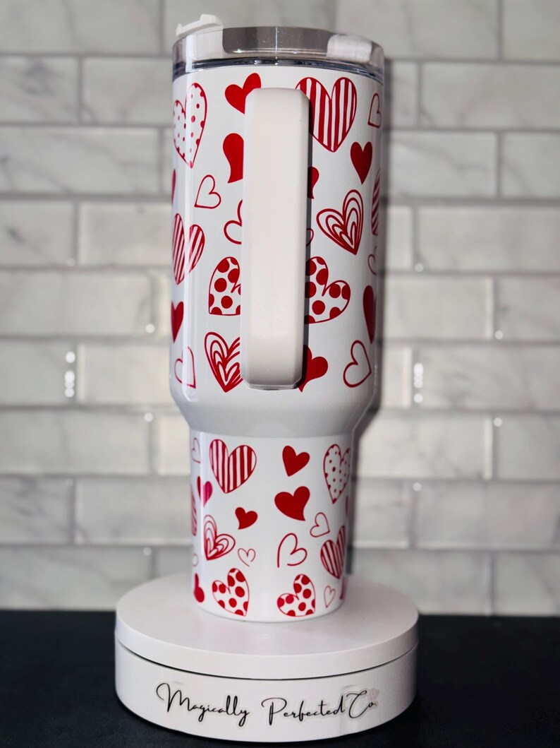 Puede incluir: Taza de viaje blanca con asa blanca y un patr&oacute;n de corazones rojos. La taza est&aacute; sobre una plataforma giratoria blanca con el texto "Magically Perfected Co."