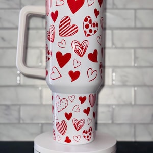 Puede incluir: Taza de viaje blanca con un patr&oacute;n de corazones rojos. La taza tiene un asa y una tapa. La taza est&aacute; sobre una plataforma giratoria blanca con el texto "Magically Perfected Co."