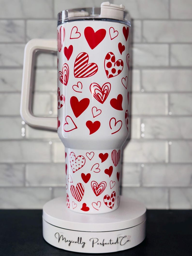 Puede incluir: Taza de viaje blanca con un patr&oacute;n de corazones rojos. La taza tiene un asa y una tapa. La taza est&aacute; sobre una plataforma giratoria blanca con el texto "Magically Perfected Co."