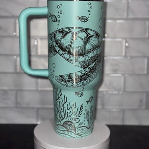 Może przedstawiać: Turkusowy kubek podróżny ze stali nierdzewnej z czarno-srebrnym wzorem przedstawiającym żółwia morskiego, koralowce i ryby. Kubek ma uchwyt i białą pokrywkę. Kubek stoi na białym cokole z nadrukiem "Magically Perfected".