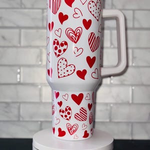 Puede incluir: Taza de viaje blanca con un patr&oacute;n de corazones rojos. La taza tiene una tapa y un asa. La taza est&aacute; sobre una plataforma giratoria blanca con el texto "Magically Perfected Co."
