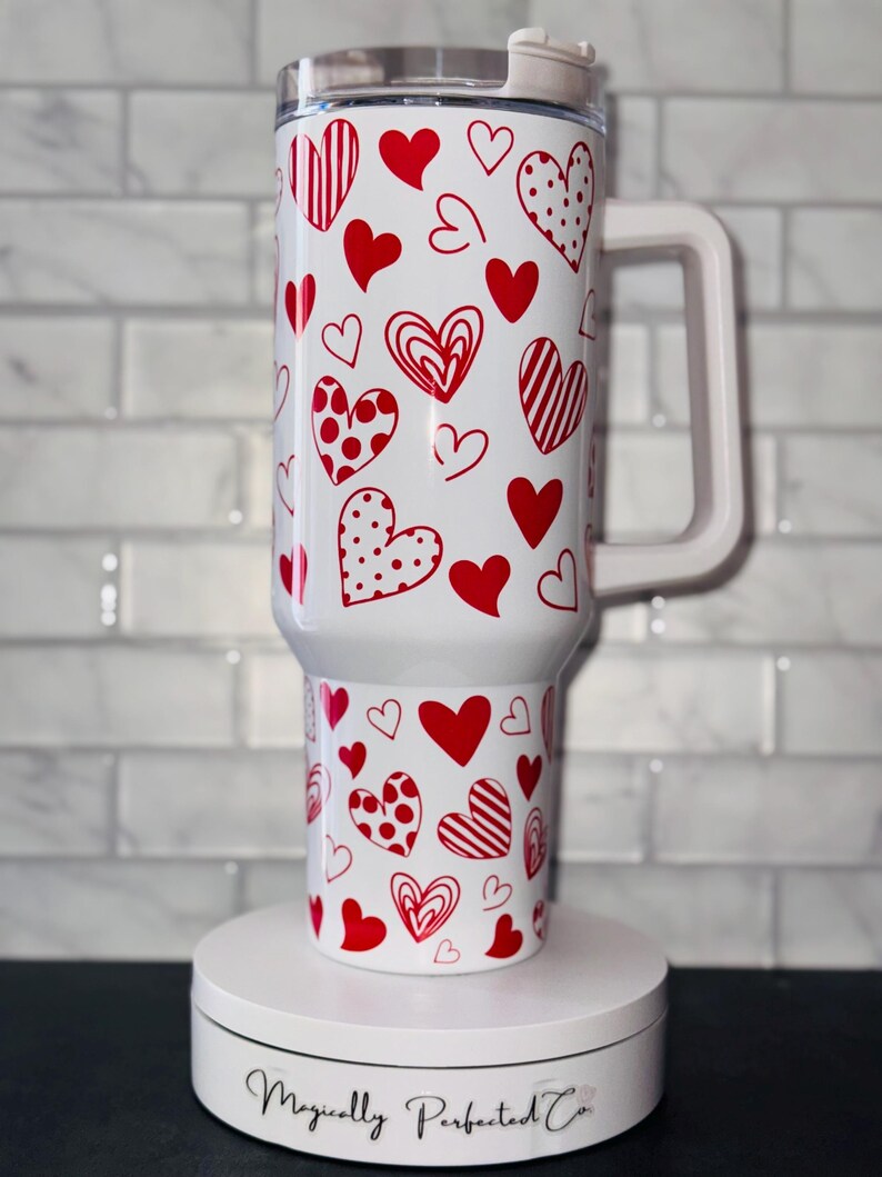 Puede incluir: Taza de viaje blanca con un patr&oacute;n de corazones rojos. La taza tiene una tapa y un asa. La taza est&aacute; sobre una plataforma giratoria blanca con el texto "Magically Perfected Co."