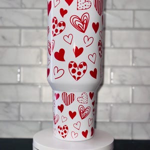 Puede incluir: Un vaso blanco con un patr&oacute;n de corazones rojos. El vaso tiene una tapa y est&aacute; sobre una plataforma giratoria blanca con el texto "Magically Perfected Co."