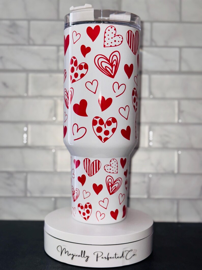 Puede incluir: Un vaso blanco con un patr&oacute;n de corazones rojos. El vaso tiene una tapa y est&aacute; sobre una plataforma giratoria blanca con el texto "Magically Perfected Co."