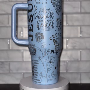 Może przedstawiać: Jasnoniebieski kubek ze stali nierdzewnej z czarnym uchwytem. Kubek ma kwiatowy wzór i inspirujące cytaty, w tym "Jezus", "ukochany", "przez jego rany zostaliśmy uzdrowieni", "stworzony z celem" i "wzrastający w łasce".