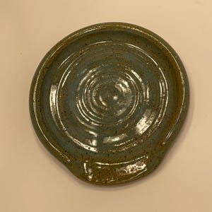 May include: A handmade ceramic spoon rest. The spoon rest is round with a shallow dish and a small lip for the spoon handle. It features a speckled blue-green glaze with concentric circles and a brown rim.
