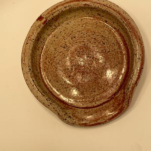 May include: A handmade ceramic spoon rest. The spoon rest is round with a small indentation for the spoon. The color is speckled tan with red and black accents. The rim is a darker shade of red.