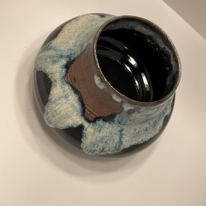 Può includere: Una ciotola in ceramica fatta a mano con interno scuro e base ampia e arrotondata. L'esterno presenta una base scura con accenti di smalto blu e bianco e una macchia marrone. L'apertura della ciotola è circolare.
