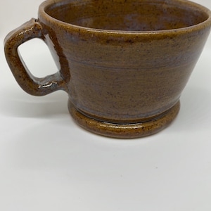 May include: A handmade brown ceramic shaving bowl with a handle. The bowl has a wide opening and a rounded base. The exterior has a textured appearance with subtle blue accents. The handle is sturdy and integrated into the bowl's design.