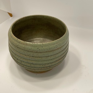 May include: A handmade, round ceramic bowl with a light green glaze and horizontal ribbed texture. The bowl has a speckled appearance and a slightly flared rim. The base is a lighter, earthy tone. This pottery piece is suitable for serving or display.