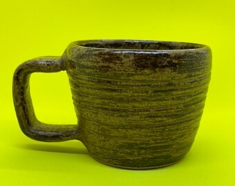 Pode incluir: Uma caneca de cerâmica artesanal com um esmalte texturizado marrom e verde terroso. A caneca apresenta uma alça quadrada e detalhes horizontais canelados ao redor do corpo. A caneca está sobre um fundo amarelo brilhante.