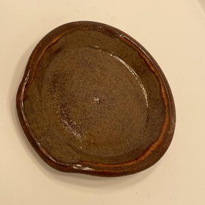 May include: A handmade, brown ceramic dish with an irregular shape. The dish has a textured surface and a darker brown rim, giving it a rustic appearance. This small dish is ideal for holding jewelry or small items, such as keys.