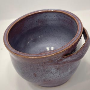 May include: A handmade ceramic bowl with a deep purple glaze and a light blue interior. The bowl has a rounded shape with a handle and a golden-brown rim. This unique piece is perfect for serving soups or stews.