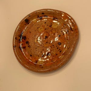 May include: A handmade ceramic plate with a speckled, organic design. The plate features a warm colour palette of brown, orange, and yellow, with dark speckles throughout. The rim is slightly raised, and the shape is irregular.