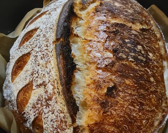 May include: A freshly baked loaf of sourdough bread with a crispy crust and a light dusting of flour. The bread is in a black cast iron pot.