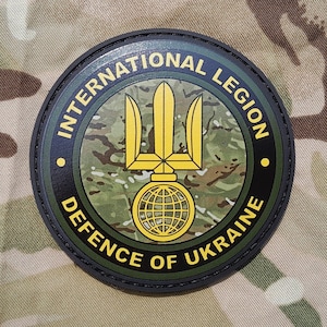 Puede incluir: Un parche redondo, negro y verde con las palabras "International Legion Defence of Ukraine" que rodean un símbolo de tridente amarillo con un globo en la base.