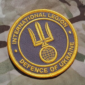 May include: A circular patch with the text "INTERNATIONAL LEGION DEFENCE OF UKRAINE" in gold thread. The patch features a gold trident and a globe design on a dark blue background, with a gold border. The patch is set against a camouflage background.