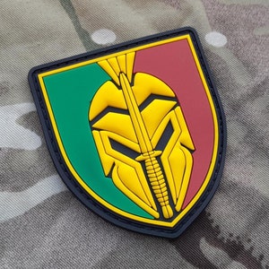 May include: A yellow and black embroidered patch with a stylized warrior's helmet and a dagger. The patch is on a green, red, and black shield shape.