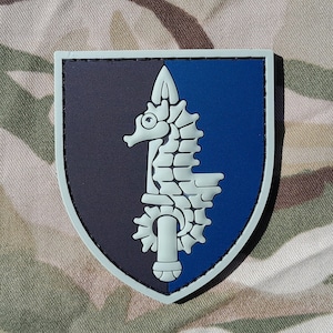 May include: A gray, blue, and white embroidered patch with a seahorse design. The patch is shaped like a shield and has a white seahorse with a black eye on a blue background. The seahorse is holding a sword in its mouth. The patch is surrounded by a gray border.