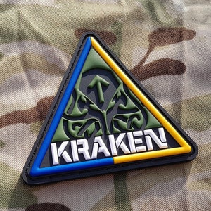 May include: A black triangular patch with a green and white symbol and the word "KRAKEN" in white letters. The patch has a blue and yellow border.
