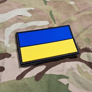 May include: A blue and yellow Ukrainian flag patch with black stitching around the edges. The patch is on a camouflage fabric background.