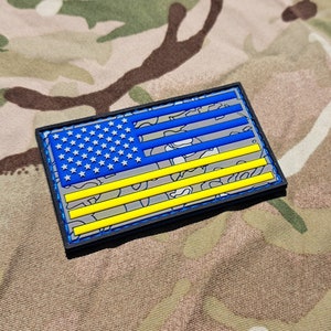 以下が含まれることがあります： 迷彩柄の青と黄色のアメリカ国旗のパッチ。パッチは耐久性のある素材で作られており、黒い縁取りがあります。