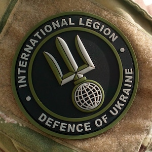 以下が含まれることがあります： 緑、黒、白のワッペンで、"International Legion Defence of Ukraine"とトライデントのシンボルが書かれています。ワッペンは迷彩柄の生地に縫い付けられています。