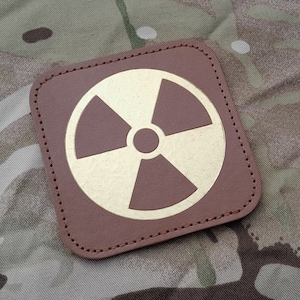 May include: A brown leather patch with a gold-colored radiation symbol. The patch has a stitched border.