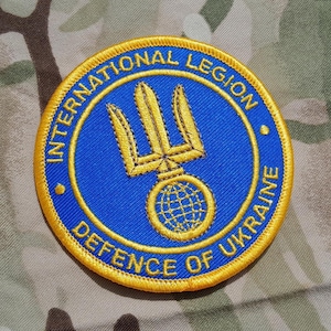 Può includere: Toppa rotonda ricamata con la scritta "INTERNATIONAL LEGION DEFENCE OF UKRAINE". La toppa presenta uno sfondo blu con un emblema di tridente e globo dorato e un bordo dorato. La toppa è su uno sfondo mimetico.