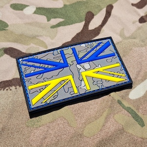Pode incluir: Um remendo de bandeira Union Jack azul e amarelo com um padrão de camuflagem. O remendo é feito de tecido e tem uma borda preta.