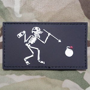 May include: A black and white embroidered patch featuring a skeleton holding a bottle and a spear, throwing a bomb. The patch has a black background and white stitching.