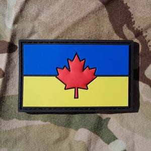 May include: A rectangular patch featuring the Canadian flag with a red maple leaf on a blue and yellow background, framed by a black border. The patch is set against a camouflage pattern.
