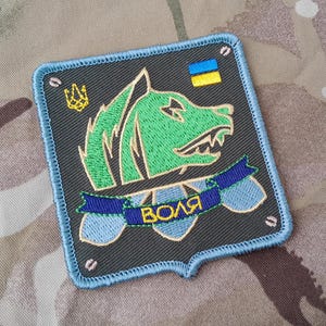 Puede incluir: Un parche bordado con la cabeza de un lobo verde y una cinta azul que dice "ВОЛЯ" en letras amarillas. El parche tiene un borde azul y una bandera ucraniana amarilla y azul en la esquina superior derecha.