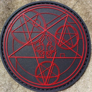 Op de afbeelding: Een ronde zwarte en rode patch met een geometrisch ontwerp. Het ontwerp heeft een grote cirkel met een vijfpuntige ster erin, omringd door kleinere cirkels en lijnen.