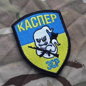 May include: A black and yellow embroidered patch featuring a ghost holding a rifle. The patch reads "KASPER" and "ЗСР" in Cyrillic.