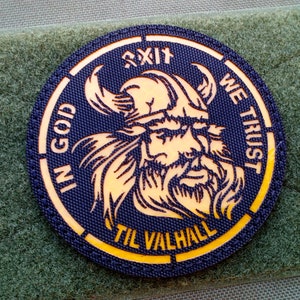 May include: A round, blue patch with a yellow border. The patch features a Viking warrior with a beard and helmet. The text "IN GOD WE TRUST" is written around the warrior's head. The text "TIL VALHALL" is written below the warrior's head.