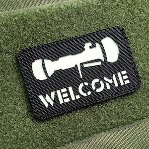 May include: A black patch with white stitching around the edges. The patch features a white silhouette of a rocket launcher and the word "WELCOME" in white letters.