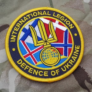 May include: A yellow embroidered patch with the text "International Legion Defence of Ukraine" surrounding a blue and yellow trident with a globe in the center. The trident is surrounded by flags of different countries.