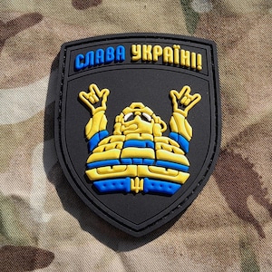 Puede incluir: Un parche negro con una figura de dibujos animados amarilla y azul con el texto "СЛАВА УКРАЇНІ!" en azul.