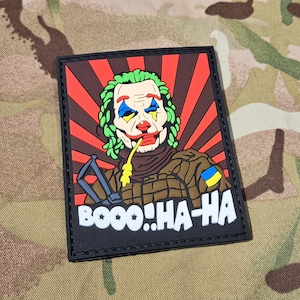 Pode incluir: Remendo retangular com um desenho de Joker de desenho animado. O Joker tem cabelo verde, maquiagem de palhaço e um uniforme marrom em estilo militar. O fundo tem raios vermelhos e marrons. O texto "BOOO! HA-HA" está na parte inferior.