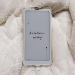 Könnte beinhalten: Ein weißer E-Reader mit einem grauen Bildschirm, auf dem der Text "I'd rather be reading" mit zwei kleinen Sternen angezeigt wird. Der E-Reader befindet sich in einem durchsichtigen Etui und liegt auf einer weißen, flauschigen Oberfläche.