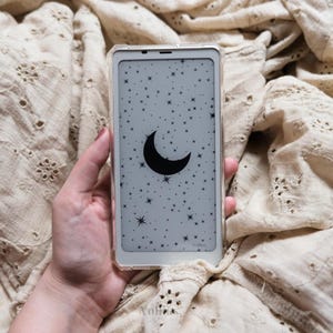 May include: A white smartphone case with a clear back showing a black crescent moon and white stars on a gray background.