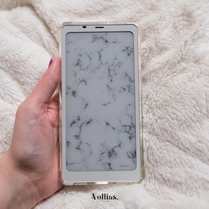 Könnte beinhalten: Eine weiße Handyhülle mit Marmormuster. Die Hülle hat eine transparente Rückseite und einen weißen Rahmen. Die Handyhülle trägt den Text "Kollias" in der unteren rechten Ecke.