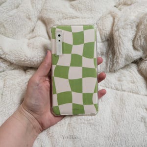 Puede incluir: Una funda transparente para teléfono con un patrón de cuadros verdes y blancos. La funda tiene una protuberancia de cámara elevada y un pequeño logotipo que dice "Yolltas".