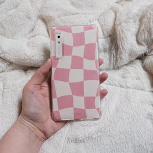 May include: A pink and white checkered phone case with a wavy pattern. The case is made of clear plastic and has a raised edge around the camera lens.