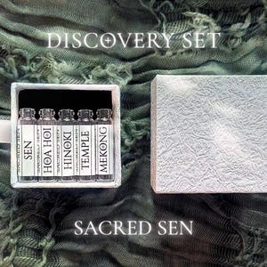 May include: A white box set labeled "DISCOVERY SET" containing five small glass bottles with white labels. The labels read "SEN", "HOA HOI", "HINOKI", "TEMPLE", and "MEKONG". The box is open, revealing the bottles inside. The words "SACRED SEN" are below the box.